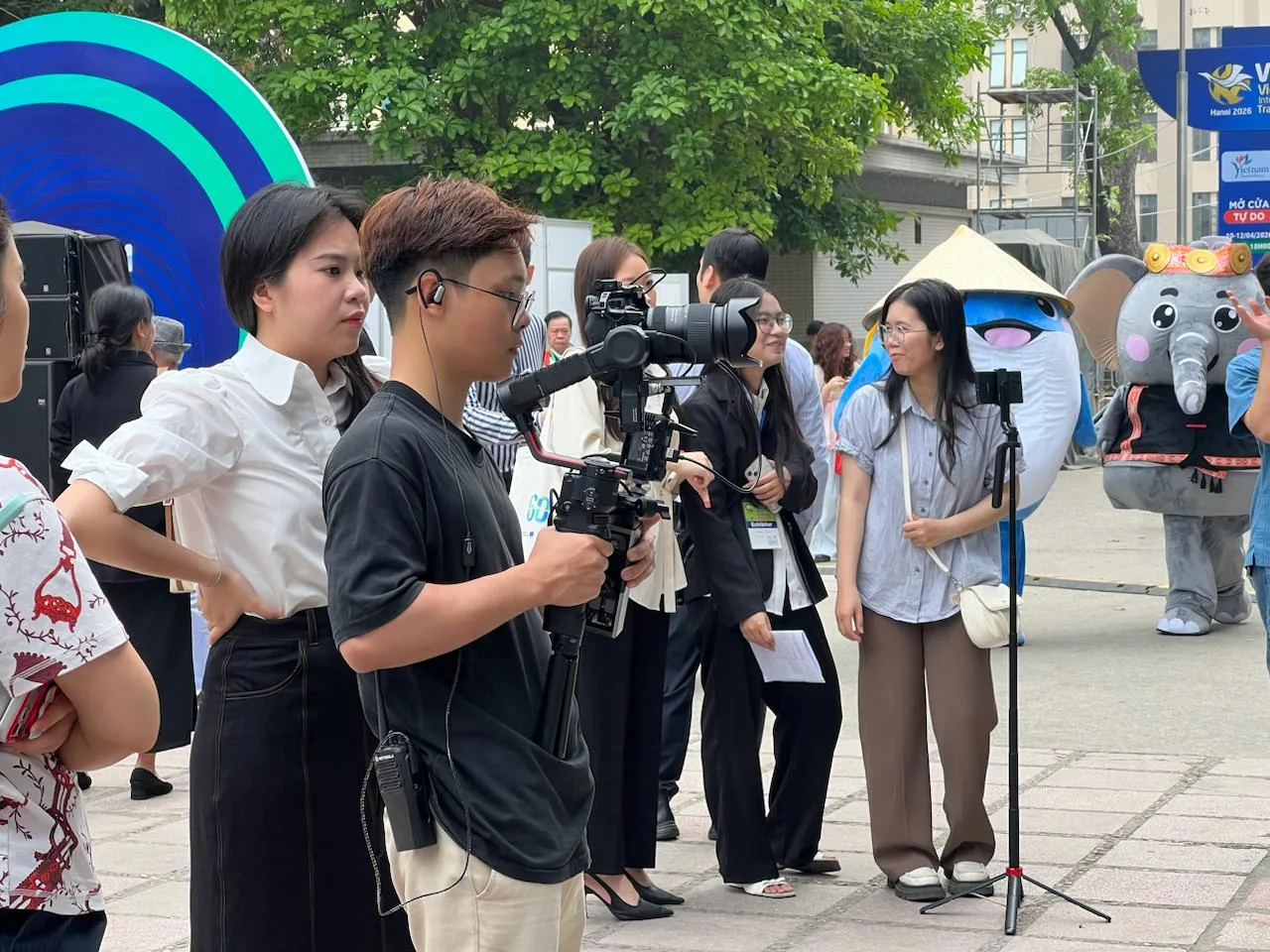 Ekip ProMedia đang quay phim hậu trường, với cameraman và các thành viên hỗ trợ, tại một sự kiện triển lãm đông người.