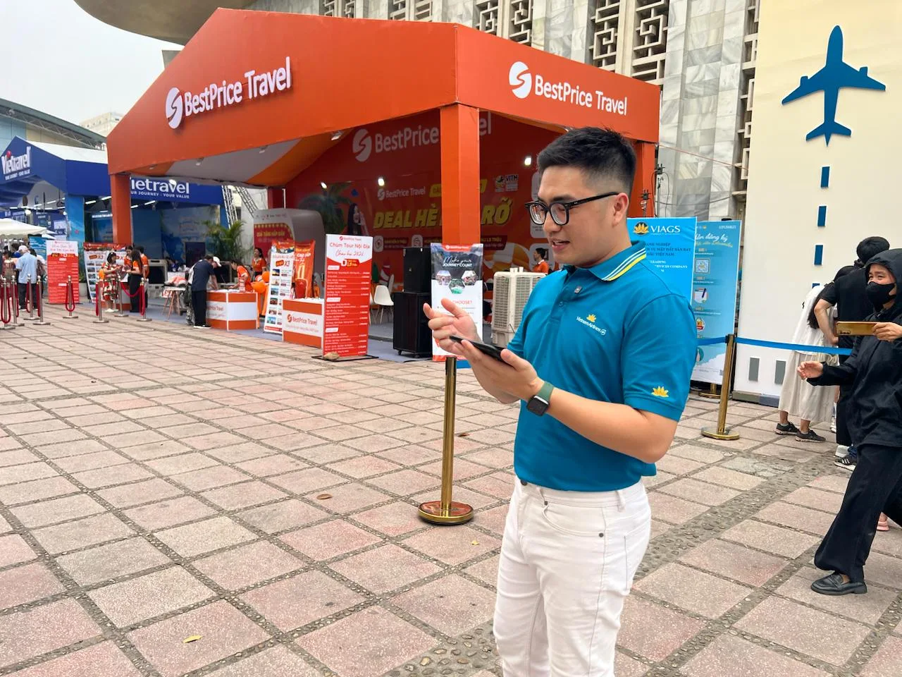 MC nam của Vietnam Airlines áo xanh đang xem điện thoại, chuẩn bị trước buổi livestream tại gian hàng BestPrice Travel.