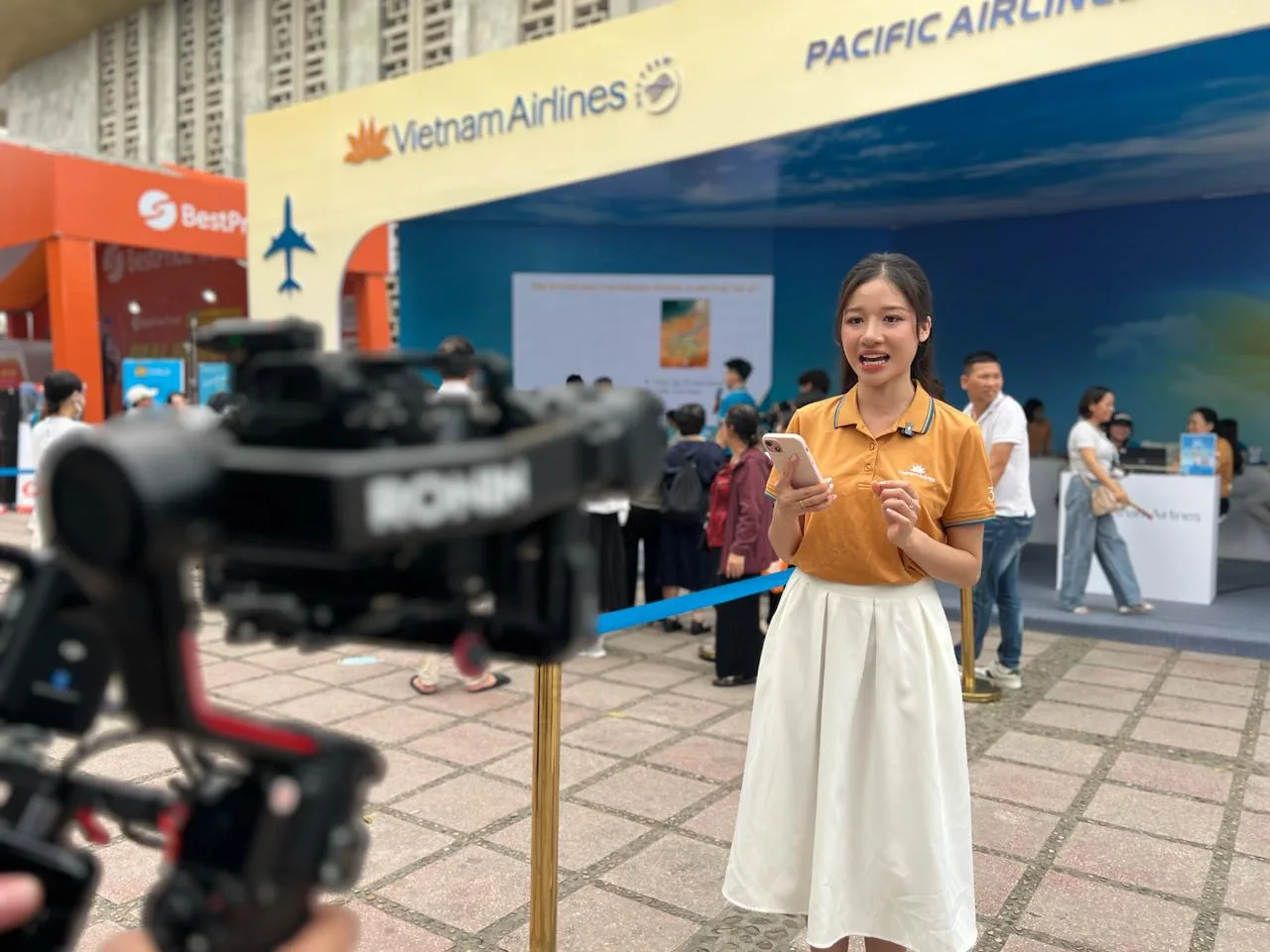 MC nữ của Vietnam Airlines áo vàng tươi cười, đang được quay hình bằng máy ảnh chuyên nghiệp với gimbal Ronin.