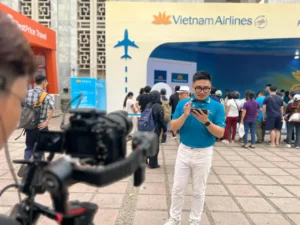 Một cameraman dùng gimbal Ronin ghi hình MC nam áo xanh của Vietnam Airlines đang tương tác với điện thoại tại triển lãm du lịch 2026.