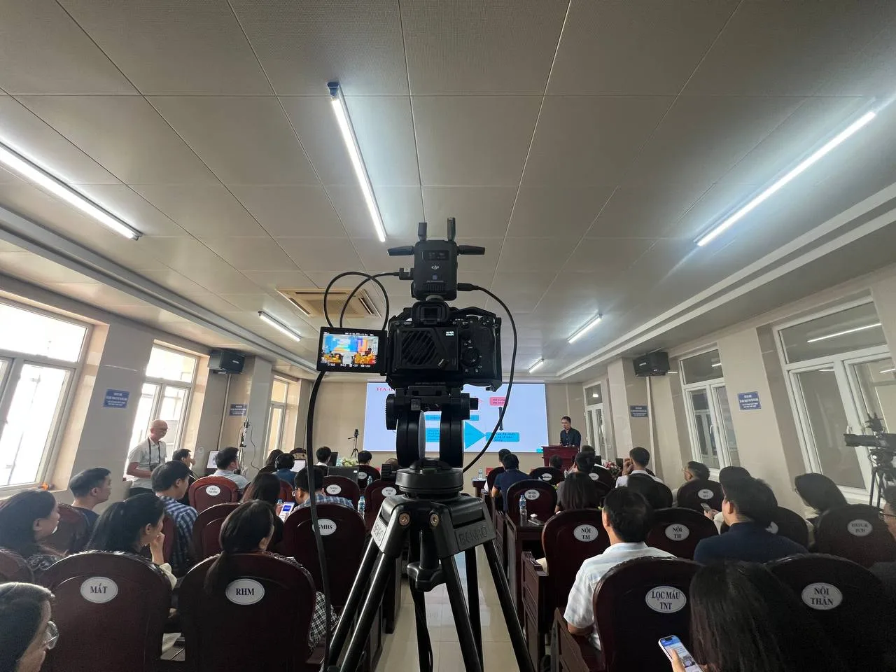 Toàn cảnh hội trường với máy quay ProMedia đang hoạt động, diễn giả trình bày.